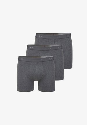 Athletischer Mann steht barfuß da und trägt dunkelgraue Boxershorts vor einem einfarbigen hellen Hintergrund.