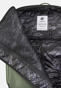 Sac à dos vert ouvert montrant l'intérieur noir avec une poche en filet zippée et une étiquette blanche de la marque Mammut à l'intérieur.
