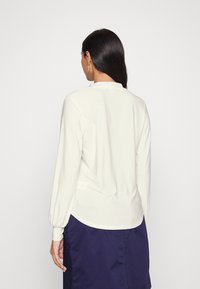 Bílá blůza s dlouhými rukávy a hladkým povrchem, mírně zakřiveným lemem a nabíranými manžetami. Zadní pohled odhaluje minimalistický design.