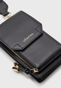 Czarna skórzana torebka na ramię z złotym zamkiem błyskawicznym, sprzętem zapięcia i wytłoczonym logo "HÉROÏNE" z przodu. Gładka faktura i zorganizowany design.