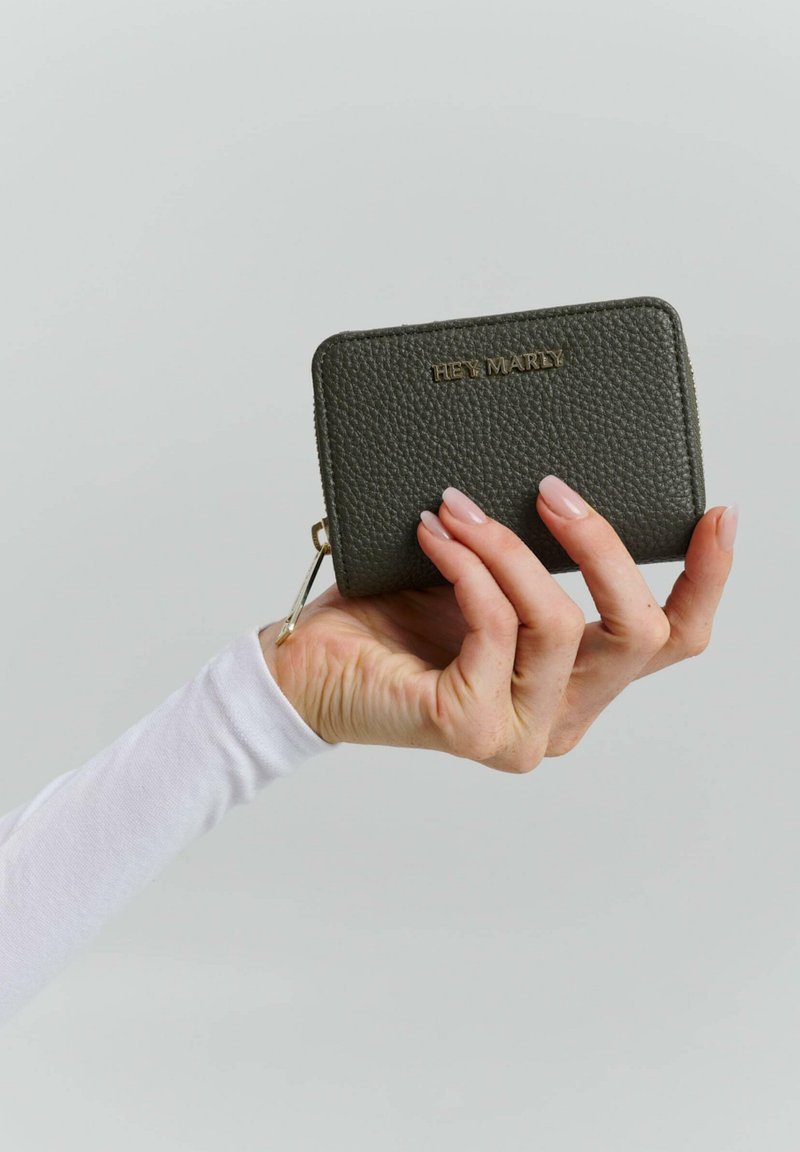 Portefeuille en cuir texturé vert foncé avec un logo doré en relief, fermeture zippée et attache pour clé, tenu par une main dans une manche blanche.