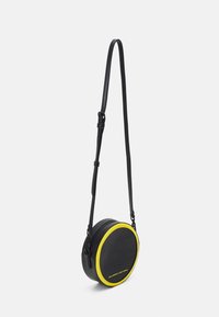 Kulatá černá crossbody taška s žlutým akcentem, vyrobená z hladkého materiálu. Má zipové zavírání a nastavitelné černé ramenní popruhy.