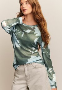 Ung kvinna med långt rödaktigt hår, klädd i en grön blommig långärmad skjorta och ljusbeige högmidjade byxor, poserar med en hand nära nacken.