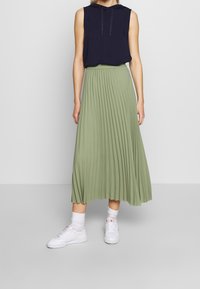 Jupe midi plissée verte, taille élastique, associée à un haut sans manches bleu marine. Des baskets blanches avec des chaussettes mi-mollet complètent la tenue.
