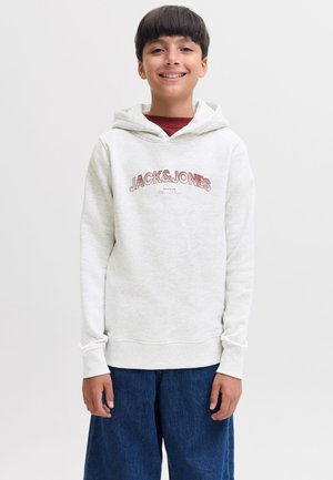 Garçon souriant portant un sweat à capuche gris clair avec le texte "JACK & JONES" et un jean bleu à jambes larges, debout devant un fond blanc uni.