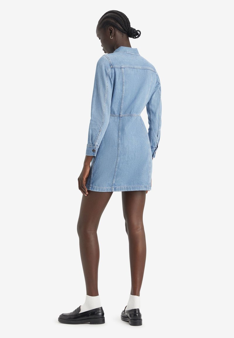 Robe chemise en denim, bleu clair, taille ajustée, poignets boutonnés, col, longueur mi-cuisse, texture lisse, design uni, portée avec des chaussures noires.