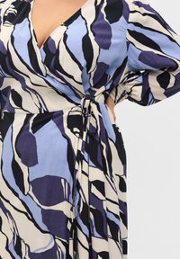 KietaisemDress, joka esittelee rohkeita sinisen, mustan ja kermanväristen abstraktien kuvioiden sekoitusta. Pehmeä kangas, jossa on pitkät hihat ja sidottu vyötäröyksityiskohta.