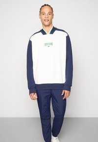 Bijeli i mornarski Reebok dukserica s zelenim logotipom, sa ovratnikom, dugim rukavima i rebrastim manžetama; uparen s mornarskim trenirka hlačama.