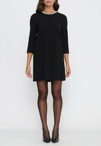 Schwarzes Kleid mit dreiviertel Ärmeln, plissiertem Vorderteil und silbernem Akzent am Ausschnitt. Getragen mit schwarzen High Heels und transparenten Strumpfhosen.