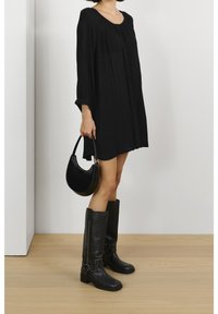 Robe noire à manches longues avec un col rond, associée à des bottes en cuir noires jusqu'aux genoux et à un sac à main noir. Tissu lisse, coupe décontractée.
