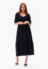 Schwarzes Maxikleid mit kurzen Ärmeln, einem Rundhalsausschnitt und einem gestuften Saum. Glattes Material mit lockerer Passform. Getragen mit schwarzen High Heels.