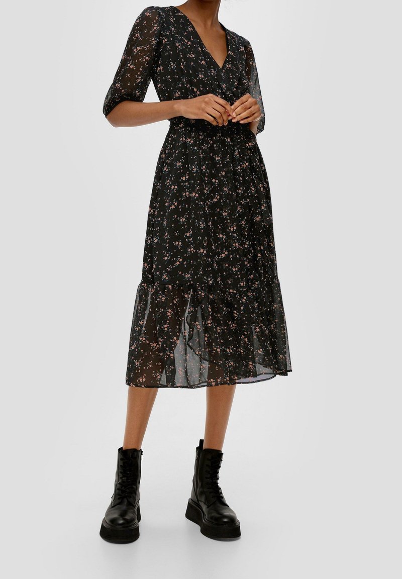 Kvinna i en svart blommig midiklänning med puffärmar, i kombination med svarta snörda kängor, står mot en enfärgad bakgrund.