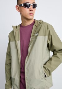 Veste à capuche légère bicolore en vert olive et beige, avec une fermeture éclair intégrale, des poches latérales et un petit logo sur la poitrine.