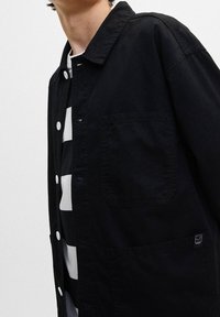 Schwarze Button-Down-Jacke aus Baumwolle mit spitzem Kragen, frontale Tasche und strukturierter Oberfläche, über einem gestreiften schwarz-weißen Hemd getragen.