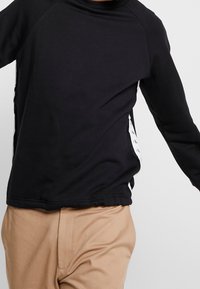 Sudadera negra de manga larga con cuello redondo, con acentos laterales blancos y una textura suave. Combinada con pantalones beige claros.