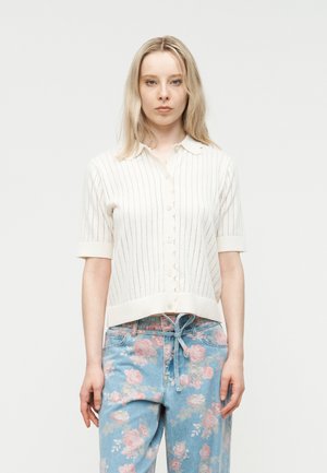 Donna bionda che indossa una maglia bianca a maniche corte con bottoni e jeans blu con motivo floreale rosa su uno sfondo bianco uniforme.