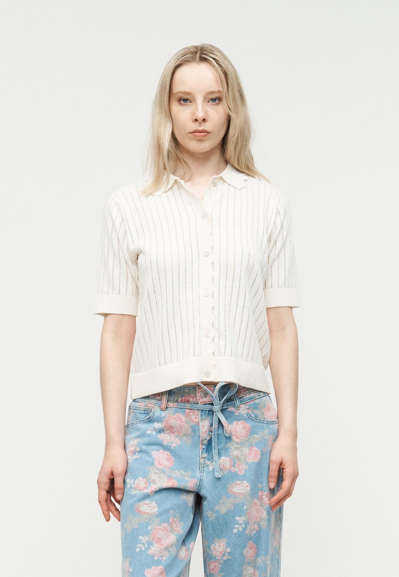 Ξανθιά γυναίκα φοράει λευκό κοντομάνικο πουκάμισο με κουμπιά και μπλε τζιν με ροζ ανθισμένο μοτίβο σε απλό λευκό φόντο.