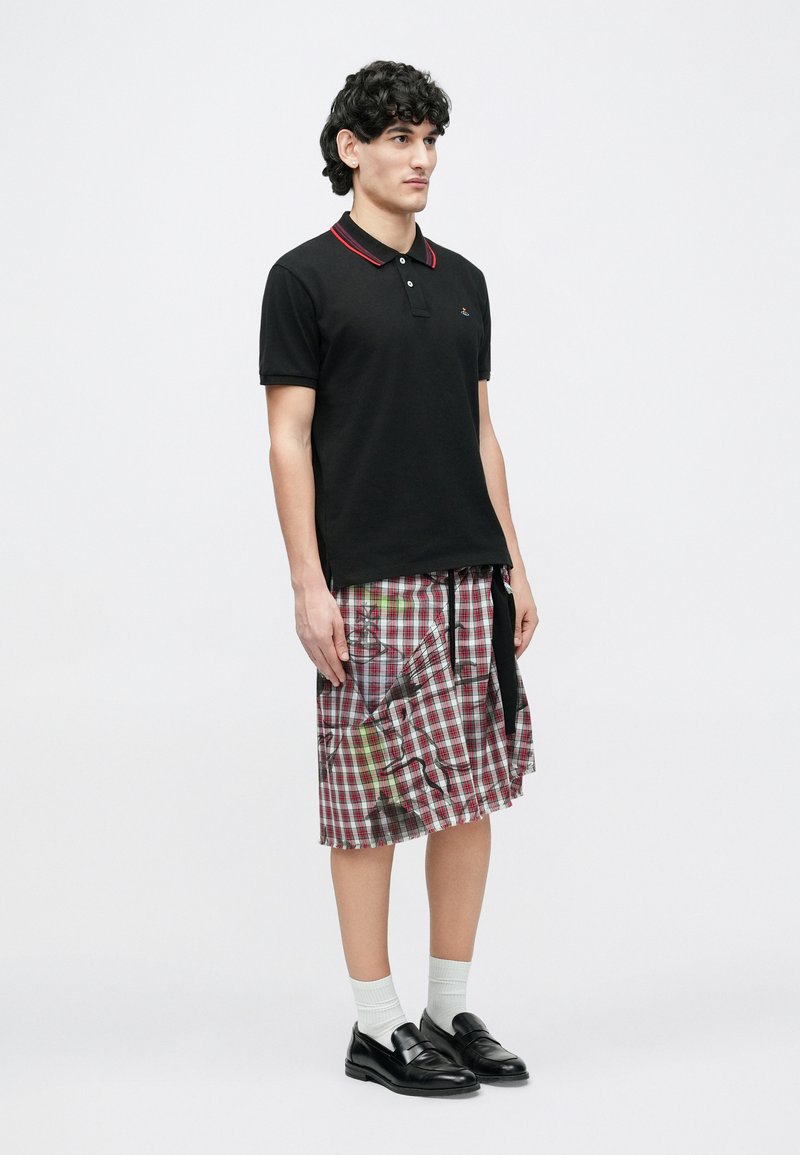 Ung mand iført en sort polo, ternet nederdel med malingssprøjt, hvide sokker og sorte loafers, stående mod en ensfarvet baggrund.