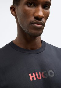 Črna bombažna majica s potiskom besede "HUGO" v rdeče-sivem prelivu. Klasičen okrogel izrez z gladko teksturo.