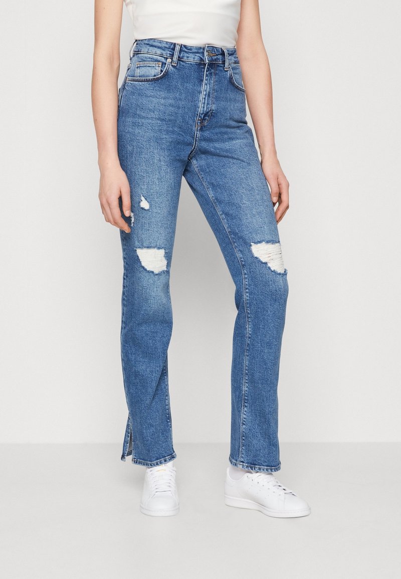 Personne portant un jean bleu déchiré taille haute avec des fentes à l'avant et des baskets blanches, debout devant un fond clair uni.