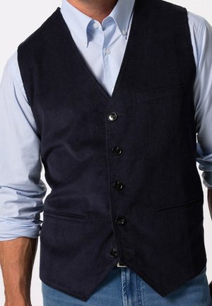 Man draagt een donkerblauwe ribfluwelen vest met knopen over een lichtblauw gestreept overhemd met opgerolde mouwen en een blauwe jeans.