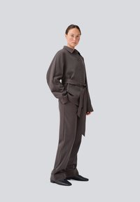 Outfit composto da camicia e pantaloni marroni su misura con cintura. La parte superiore presenta un colletto, bottoni e maniche lunghe leggermente arricciate. Tessuto morbido.