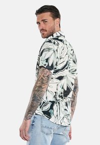 Kurzarmhemd mit einem schwarz-weißen Blumenmuster und grünen Akzenten, der Stoff hat eine glatte Textur und ist mit einer Knopfleiste gestaltet.