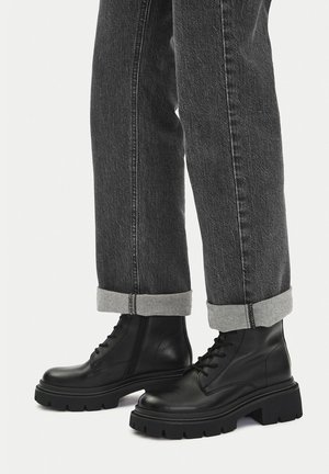 Czarne skórzane botki za kostkę z zaokrąglonym czubkiem, grubą podeszwą i sznurowaniem, zestawione z szarymi podwiniętymi dżinsami.