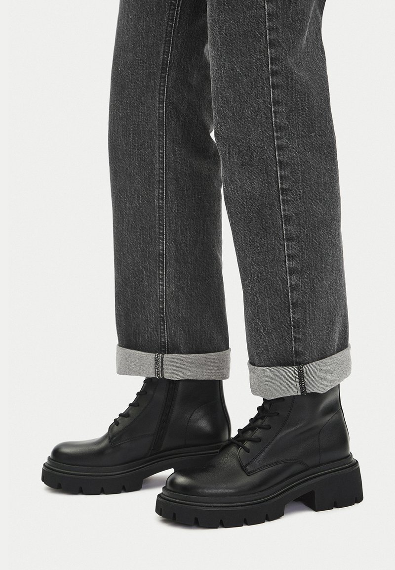 Czarne skórzane botki za kostkę z zaokrąglonym czubkiem, grubą podeszwą i sznurowaniem, zestawione z szarymi podwiniętymi dżinsami.