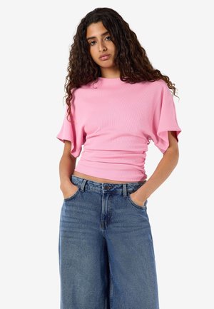 Kvinde med langt krøllet hår iført en pink kortærmet top og højtaljede, vide blå jeans med hænderne i lommerne.