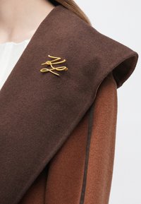 Palton maro din fetru cu un guler supradimensionat, având o broșă abstractă aurie pe rever. Material texturat în tonuri calde.