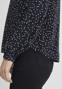 Marineblaue Bluse mit weißen Punkten, langen Ärmeln mit elastischen Bündchen, einem geschwungenen Saum und einem Knopfdetail am Rücken.