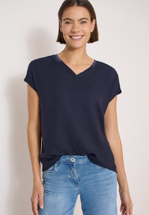 Smilende kvinde med kort brunt hår iført en mørkeblå kortærmet V-hals top og lyseblå jeans mod en ensfarvet baggrund.