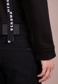 Giacca in denim nera con un'etichetta "VERSUS". Presenta una fascia bianca a contrasto, bottoni in metallo e dettagli di cuciture meticolose.