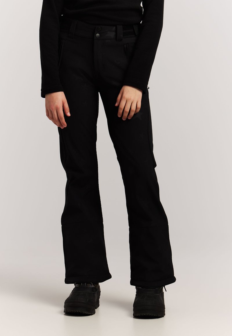 Personne portant un pantalon évasé noir, un haut à manches longues noir et des chaussures noires, se tenant devant un fond clair uni.