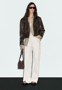 Bruine leren cropped jas, beige shirt, witte wijde broek, donkere zonnebril en bruine schoudertas. Gladde texturen en neutrale kleuren.