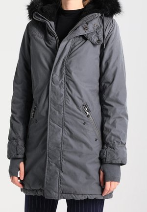 Personne portant un manteau d'hiver gris mi-long avec une capuche bordée de fourrure noire, des poches zippées et des manches de moufles sans doigts.