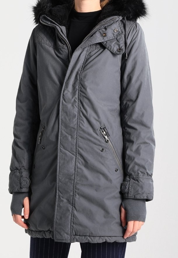 Personne portant un manteau d'hiver gris mi-long avec une capuche bordée de fourrure noire, des poches zippées et des manches de moufles sans doigts.