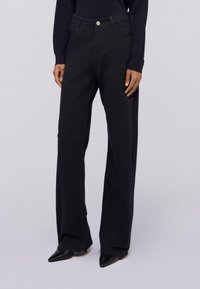 Persona vestida con jeans de tiro alto y pierna ancha negros, zapatos de punta negra y una blusa de manga larga negra, de pie contra un fondo liso.