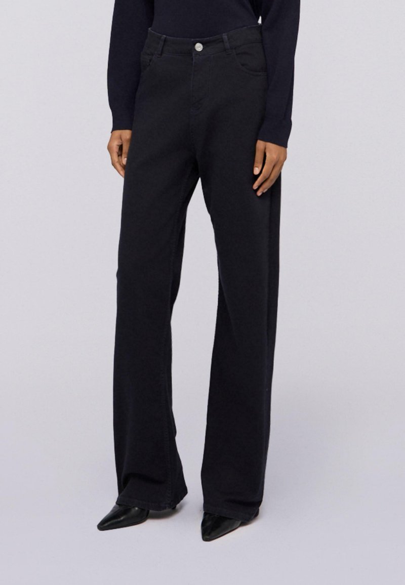 Persona vestida con jeans de tiro alto y pierna ancha negros, zapatos de punta negra y una blusa de manga larga negra, de pie contra un fondo liso.