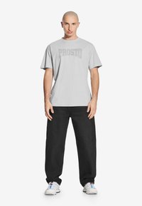 Jasnoszara koszulka z logo "PROSTO" w ciemniejszym odcieniu, połączona z luźnymi czarnymi dżinsami i białymi butami sportowymi. Teksturowany materiał, casualowy krój.