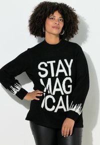 Suéter negro con texto blanco en negrita "STAY MAGICAL" y detalles texturizados blancos en las mangas. Modelo combinado con pantalones negros brillantes.