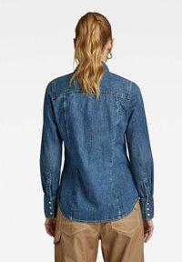 Camicia in denim blu scuro con un design aderente, polsini abbottonati e orlo curvo. Presenta dettagli di cucitura sulle spalle e sulle cuciture.
