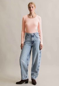 Jovem mulher em pé, vestindo uma blusa de manga longa canelada rosa claro, jeans azuis claros de cintura alta e botas de tornozelo castanhas escuras.