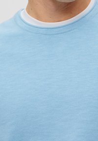 Close-up of a person wearing a light blue knit sweater over a white crew-neck shirt against a plain light background.