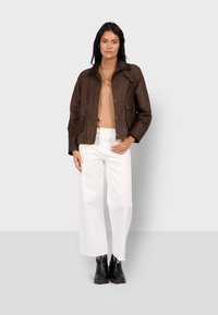 Hnědá kožená bunda s límečkem a dvěma předními kapsami, nošená přes béžový rolák a bílé kalhoty s širokými nohavicemi. Zvýrazněné černé kotníkové boty.