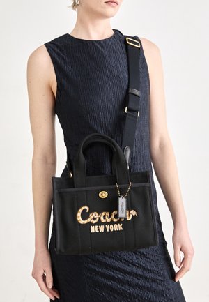 Femme portant une robe noire sans manches porte un sac cabas noir Coach New York avec une large bandoulière et des détails dorés à travers son torse.