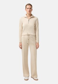 Beżowa bluzka na zamek z długim rękawem i dopasowane, wysokie spodnie z talią na sznurek oraz prostymi nogawkami, wykonane z gładkiego materiału.