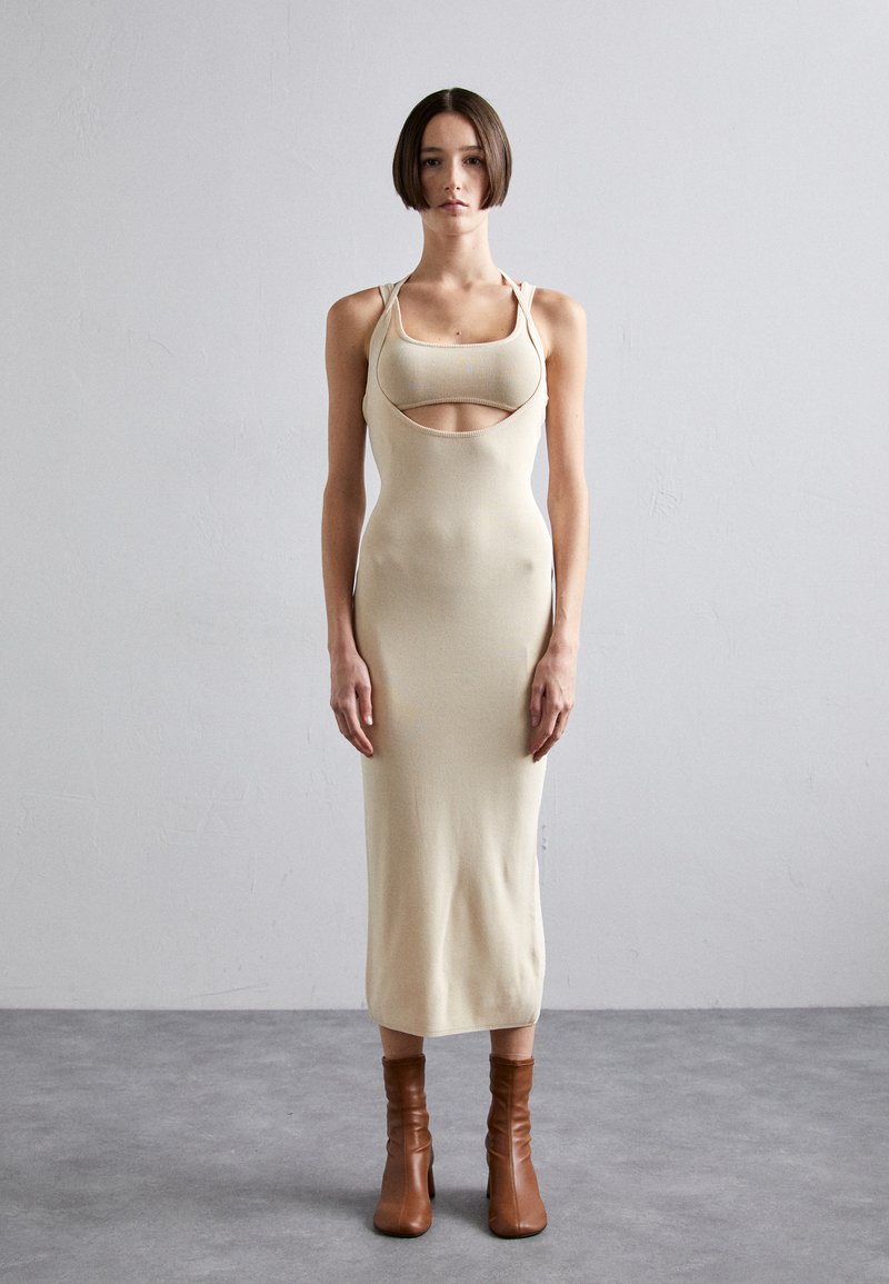 Beige fitted dress with cut-out detail and adjustable straps, paired with brown ankle boots and a smooth, textured finish.