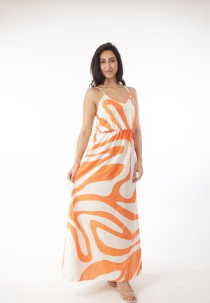 Vrouw die een mouwloze witte maxi-jurk draagt met gedurfde oranje abstracte patronen, staand en de rok vasthoudend op een witte achtergrond.